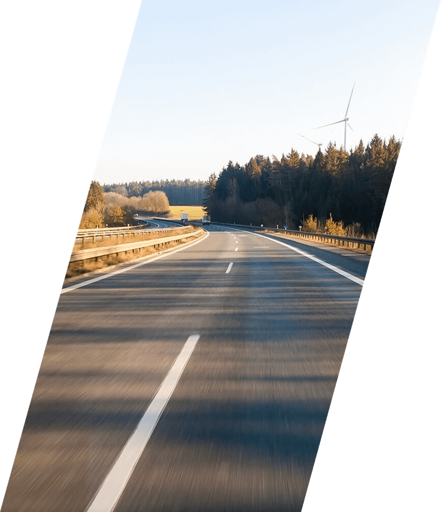 A quiet highway curves through the rural outskirts of Scunthorpe, lined by trees on both sides. In the distance, a wind turbine stands tall against a clear blue sky. The image is shown at a diagonal angle, giving a dynamic view of the peaceful countryside.